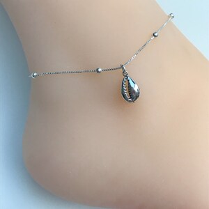 May include: A silver anklet featuring a delicate chain with small ball accents. A seashell charm hangs from the chain. The anklet is designed to be worn around the ankle.