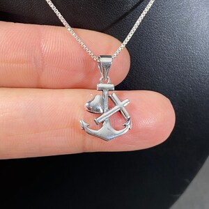 May include: A silver-colored pendant necklace featuring an anchor design with a heart and a cross. The pendant hangs from a delicate chain. The necklace is displayed against a dark background.