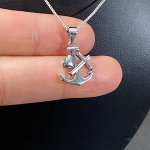 May include: A silver pendant necklace featuring an anchor design. The pendant includes a heart, a cross, and the anchor. The necklace has a delicate chain and is displayed against a dark background.
