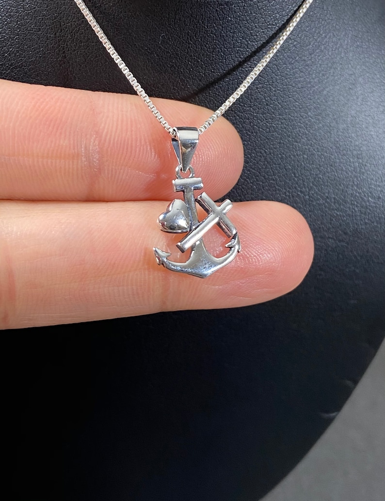 May include: A silver pendant necklace featuring an anchor design. The pendant includes a heart, a cross, and the anchor. The necklace has a delicate chain and is displayed against a dark background.