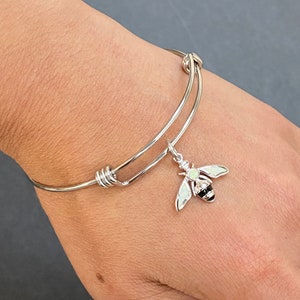 May include: Silver-toned adjustable bangle bracelet with a bee charm. The bee charm has white wings, a black and silver striped body, and a small, round, white stone. The bracelet is shown on a person's wrist.