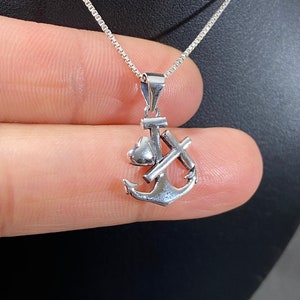 May include: A silver pendant necklace featuring an anchor, cross, and heart design. The pendant is suspended from a delicate silver chain. The necklace is displayed against a dark background.