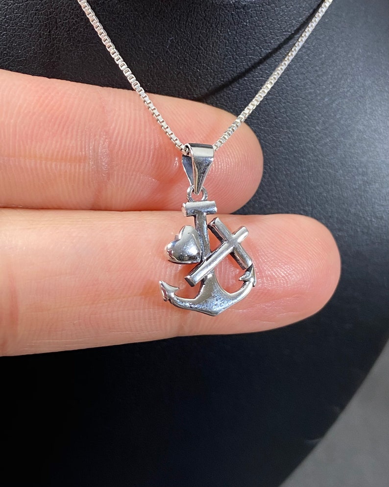 May include: A silver pendant necklace featuring an anchor, cross, and heart design. The pendant is suspended from a delicate silver chain. The necklace is displayed against a dark background.