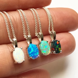 May include: Four silver necklaces with oval opal pendants in white, blue, green, and black. The opals are set in silver bezels and hang from delicate silver chains. The pendants are approximately 0.5 inches in length.
