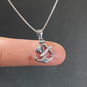 May include: A silver-colored pendant necklace featuring an anchor, a heart, and a cross. The pendant is suspended from a delicate silver chain. The necklace is shown against a dark background.