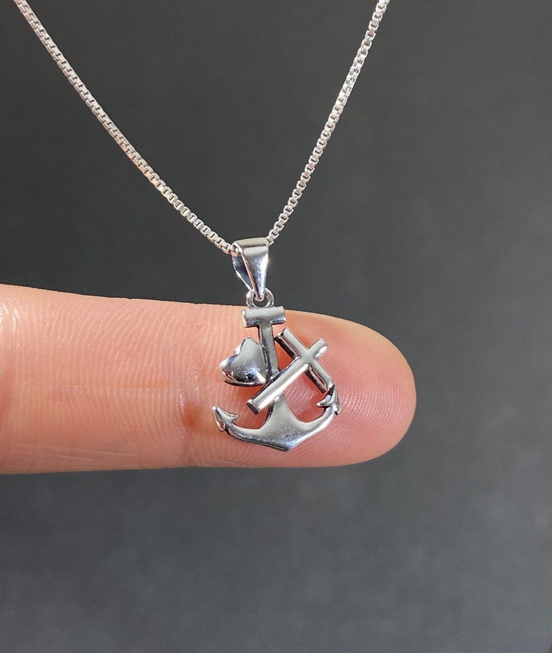 May include: A silver-colored pendant necklace featuring an anchor, a heart, and a cross. The pendant is suspended from a delicate silver chain. The necklace is shown against a dark background.