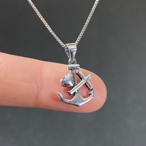 May include: A silver-colored pendant necklace featuring an anchor design with a heart and cross. The pendant hangs from a delicate chain. The anchor is detailed with a heart and cross, and the necklace is displayed against a neutral background.