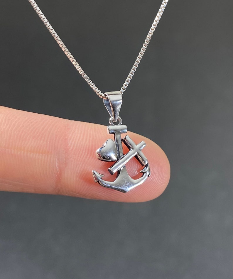 May include: A silver-colored pendant necklace featuring an anchor design with a heart and cross. The pendant hangs from a delicate chain. The anchor is detailed with a heart and cross, and the necklace is displayed against a neutral background.