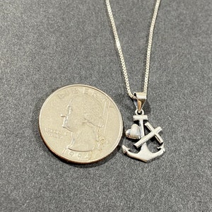May include: A silver-colored pendant necklace featuring an anchor, heart, and cross design. The necklace is displayed next to a U.S. quarter for size comparison. The chain is a delicate box chain.