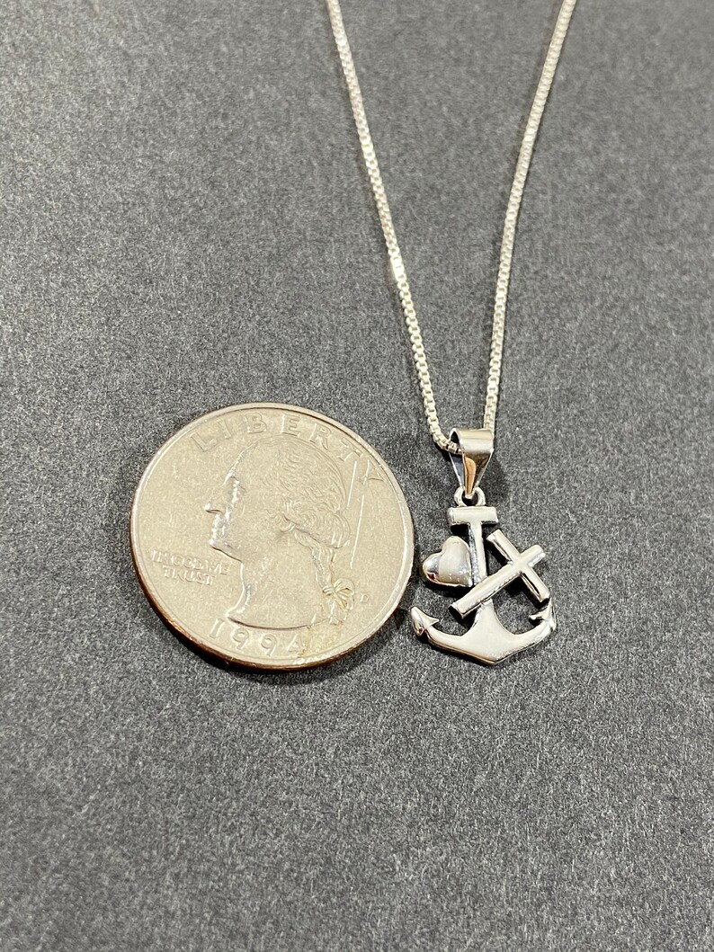 May include: A silver-colored pendant necklace featuring an anchor, heart, and cross design. The necklace is displayed next to a U.S. quarter for size comparison. The chain is a delicate box chain.
