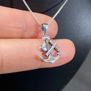 May include: A silver pendant necklace featuring an anchor, cross, and heart charm. The charm is attached to a delicate chain. The pendant is held by a person's fingers against a dark background.