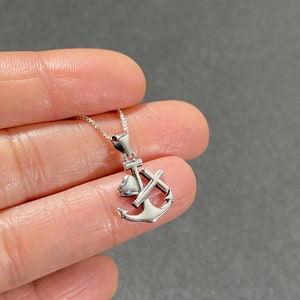 May include: A silver-colored pendant necklace featuring an anchor design with a heart and cross. The pendant is suspended from a delicate chain. The necklace is displayed against a gray background.