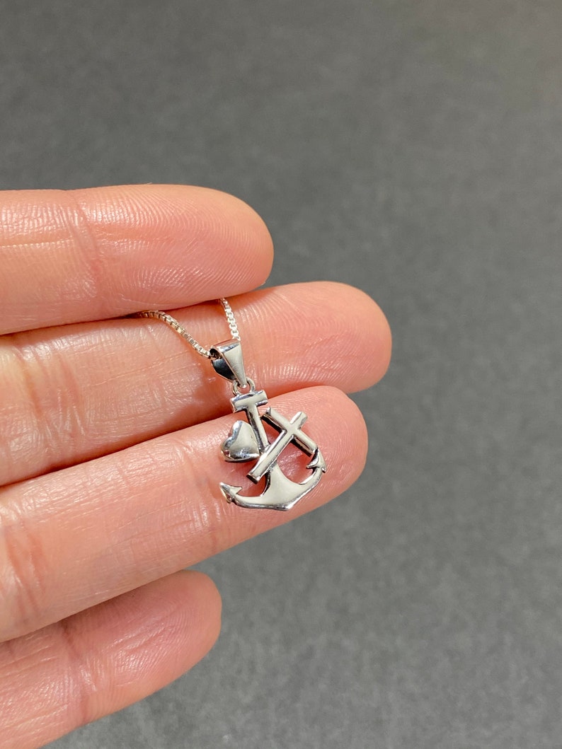 May include: A silver-colored pendant necklace featuring an anchor design with a heart and cross. The pendant is suspended from a delicate chain. The necklace is displayed against a gray background.