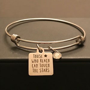 May include: Silver-toned bangle bracelet with a square charm and a small, round crystal. The charm is engraved with the words "THOSE WHO REACH CAN TOUCH THE STARS" and a star symbol. The bracelet is displayed on a dark surface.