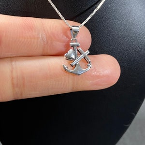 May include: A silver-colored pendant necklace featuring an anchor, heart, and cross design. The pendant is suspended from a delicate chain. The necklace is displayed against a dark background.