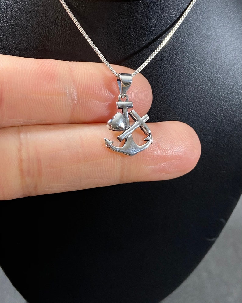 May include: A silver-colored pendant necklace featuring an anchor, heart, and cross design. The pendant is suspended from a delicate chain. The necklace is displayed against a dark background.