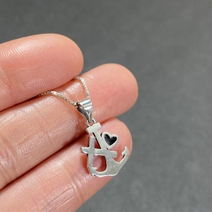 May include: A silver-colored pendant necklace featuring an anchor design with a small black heart. The pendant is attached to a delicate chain. The anchor design includes the letters "I" and "A".