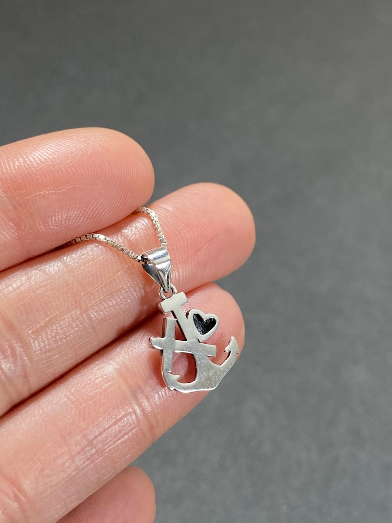 May include: A silver-colored pendant necklace featuring an anchor design with a small black heart. The pendant is attached to a delicate chain. The anchor design includes the letters "I" and "A".