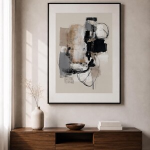 May include: A framed abstract wall art print featuring a composition of layered shapes and gestural lines in shades of black, charcoal gray, beige, and tan, set against a neutral background within a thin black frame.