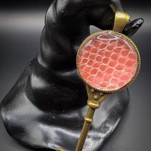 May include: A bronze key pendant with a circular glass charm. The charm displays a red and white snakeskin design. The key is set against a black, textured backdrop.
