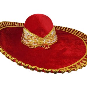 May include: A red velvet sombrero featuring a wide brim with gold metallic braided trim along the edge. The crown is accented with a white band decorated with intricate gold embroidery and a gold cord tied in a bow at the front base.