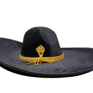 Peut inclure: Un sombrero noir avec un large bord incurvé et une calotte haute et arrondie. Le chapeau est orné d'une bordure tressée dorée qui entoure la base de la calotte et présente un détail de nœud doré décoratif sur le devant.