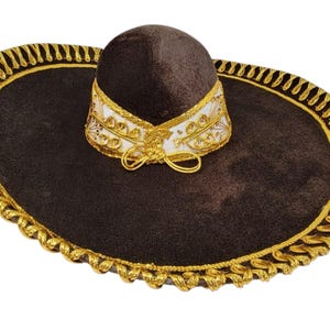 May include: A dark brown sombrero featuring a wide brim with a decorative gold braided trim along the edge. The crown is encircled by a white band adorned with intricate gold embroidery and a small gold bow detail at the front centre.