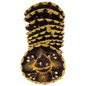 Op de afbeelding: Een stapel kleine donkerbruine sombrero-hoeden met gouden metallic randen langs de zijkanten en de kroon. Elke hoed is versierd met ingewikkeld goudkleurig borduurwerk in een zonnestraalpatroon en kleine ronde gouden accenten op de rand.