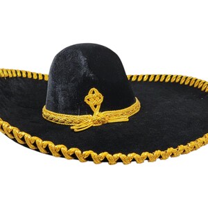 May include: A black velvet sombrero featuring a wide brim and a high crown. The hat is decorated with metallic gold braided trim along the outer edge of the brim and a matching gold braided band around the base of the crown with a knot detail.