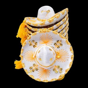 May include: A stack of white sombrero hats featuring bright yellow decorative trim, gold-coloured sequins, and yellow thread embroidery radiating from the centre. Each hat includes a yellow tassel hanging from the side of the wide brim.