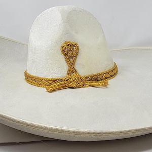 Op de afbeelding: Een witte hoed met een brede rand, een hoge, ronde kroon en een wijd uitlopende rand. De hoed is versierd met een metallic gouden gevlochten bies die de basis van de kroon omcirkelt en een decoratief knoopdetail in het midden aan de voorzijde.