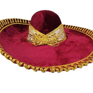May include: A deep burgundy velvet sombrero featuring a wide brim with gold metallic braided trim along the edge. The crown is accented with a white band decorated with gold embroidery and a gold cord tied in a bow at the front base.