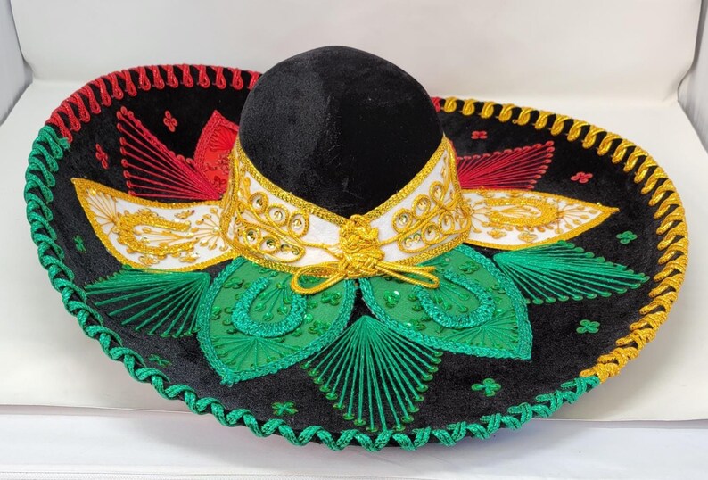 May include: A black sombrero with a rounded crown and wide, flat brim. The hat features red, green, and gold embroidered details, including leaf patterns and a decorative band around the crown. The brim is trimmed with a green and gold rope.