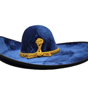 May include: A deep blue velvet sombrero featuring a wide, upturned brim with a black edge trim. The crown is adorned with a decorative gold braided cord that wraps around the base and forms a knotted detail at the front of the hat.