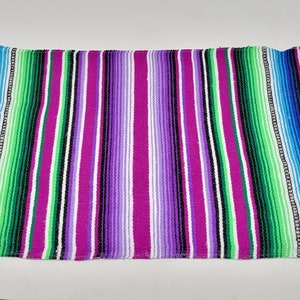 Może przedstawiać: Kolorowy meksykański koc w pasy w kolorze różowym, fioletowym, zielonym, niebieskim i białym. Koc ma białe frędzle po wszystkich stronach.