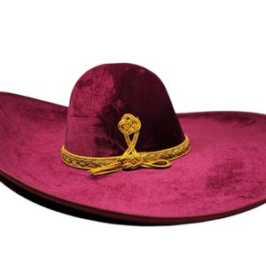 May include: A deep burgundy velvet sombrero featuring a wide, curved brim and a high, rounded crown. The hat is decorated with a gold braided trim around the base of the crown and a decorative gold knot detail centred on the front of the hat.