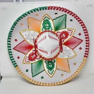 May include: A white sombrero with red, green, orange, and gold embroidered details. The hat features a circular design with a central crown and a wide, decorated brim. The brim is trimmed with green, red, and gold accents.