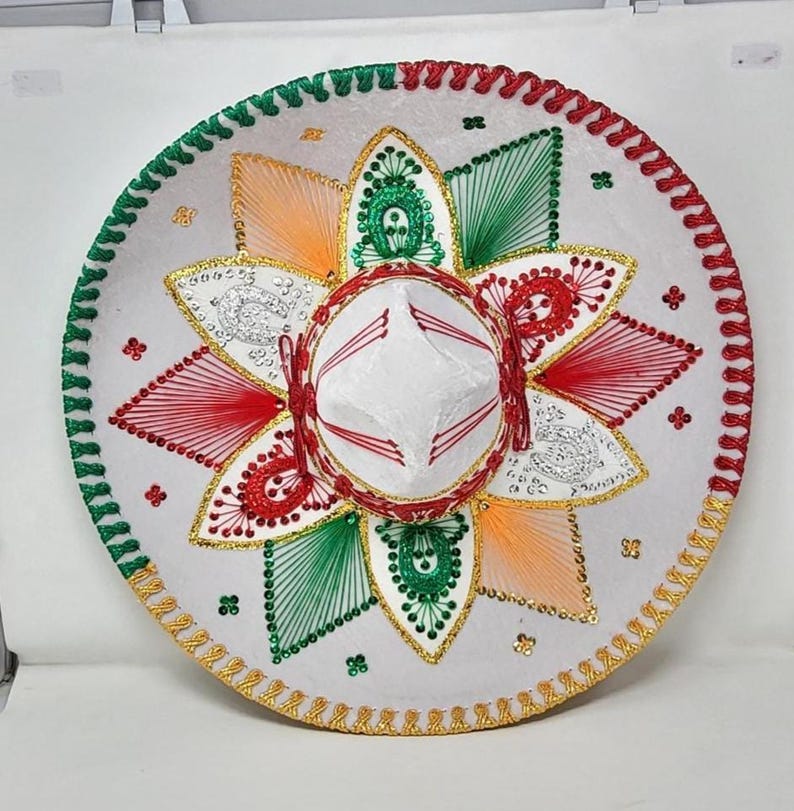 May include: A white sombrero with red, green, orange, and gold embroidered details. The hat features a circular design with a central crown and a wide, decorated brim. The brim is trimmed with green, red, and gold accents.