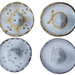 May include: Four white sombreros arranged in a grid. Two hats feature gold trim and gold embroidery with sequins, while the other two feature silver trim and silver embroidery with sequins. Each hat has a circular crown and a wide, flat brim.