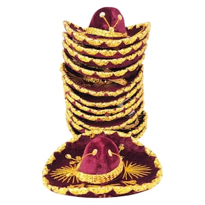 Può includere: Una pila di sombrero in velluto bordeaux scuro con finiture metalliche dorate lungo i bordi e la corona. Ogni cappello è decorato con un intricato ricamo in filo d'oro che si irradia dal centro, creando un motivo coerente sull'insieme.