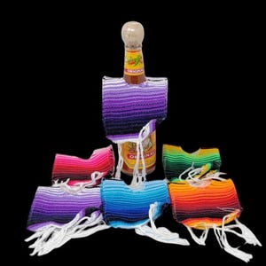 May include: A set of six colorful bottle covers with fringe. The covers are made of woven fabric and feature a variety of colors, including red, orange, yellow, green, blue, and purple. The covers are designed to fit around the neck of a bottle, such as a hot sauce bottle.