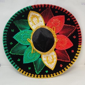 May include: A black sombrero with colorful embroidery. The hat features red, green, and white floral designs, gold trim, and a black velvet crown. The brim is decorated with gold and green stitching.