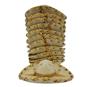 Op de afbeelding: Een verticale stapel miniatuur gebroken witte sombrero's met goudkleurige metalen randen langs de hoedenranden. Elke hoed heeft een ronde kroon en een brede rand, met gouden draaddetails rond de basis van de kroon en decoratieve gouden accenten.