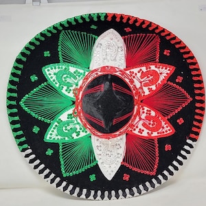 May include: A large, round sombrero in black, red, green, and white. The hat features intricate embroidery and sequin details, with a central crown and a wide brim. The design incorporates the colors of the Mexican flag.