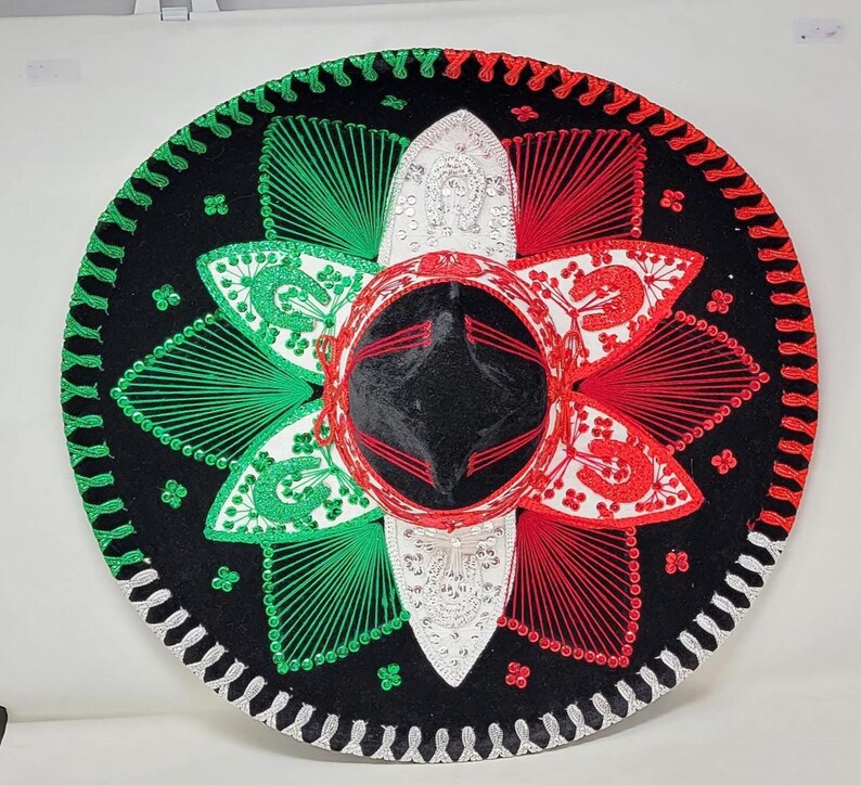 May include: A large, round sombrero in black, red, green, and white. The hat features intricate embroidery and sequin details, with a central crown and a wide brim. The design incorporates the colors of the Mexican flag.