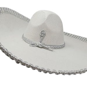 May include: A white sombrero featuring a wide, curved brim and a high, rounded crown. The hat is decorated with intricate silver metallic trim along the edge of the brim and a matching silver braided band around the base of the crown.