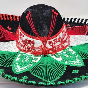 May include: A traditional sombrero with a wide brim, featuring a black crown and brim with red, white, and green embroidered details. The hat has a decorative design with a white band and red accents.