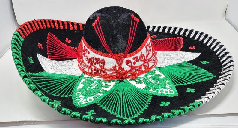 May include: A traditional sombrero with a wide brim, featuring a black crown and brim with red, white, and green embroidered details. The hat has a decorative design with a white band and red accents.