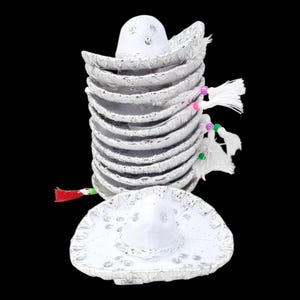 May include: A stack of white miniature sombreros with silver metallic trim and detailing. Each hat features a small white tassel attached to the side with a colourful bead. One individual white sombrero with silver trim is placed in front of the stack.