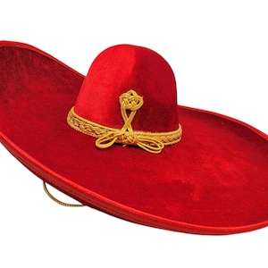 Op de afbeelding: Een felrode fluwelen sombrero met een brede rand en een hoge, ronde kroon. De hoed is versierd met een gouden gevlochten bies rond de basis van de kroon en een decoratief gouden knoopdetail aan de voorzijde, inclusief een dunne gouden kinband.